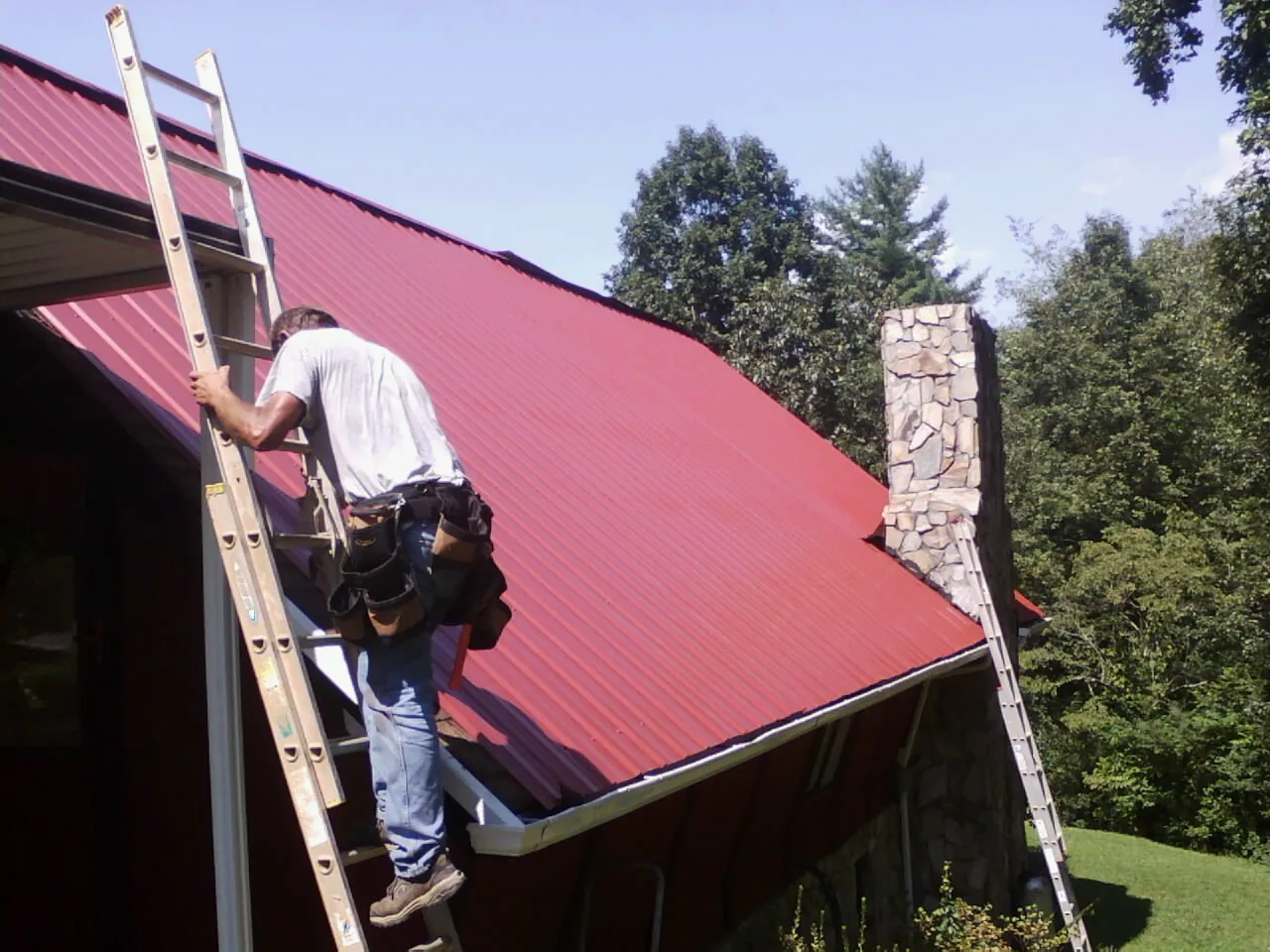 Roofer on a ladder beside a red metal roof and stone chimney — Soffit & Fascia Repair by Roofing Brothers in Stinson Beach